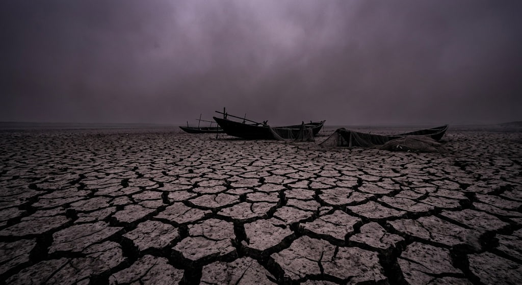 High-contrast realistic photo of a dried-up cracked mud riverbed under a dark purple and grey hazy sky causing a nuclear drought.
